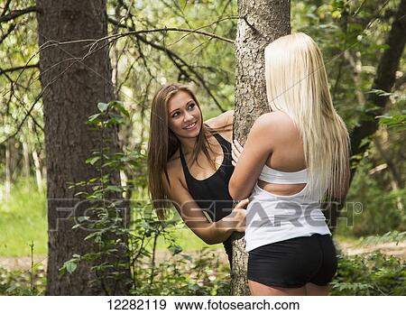 Stock Photo - One girlfriend peaking out behind a tree at another girlfriend in a park; Edmonton, Alberta, Canada. Fotosearch