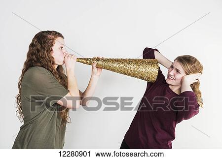 Stock Image - Studio shot of a young woman blowing loudly at her friend using a fake horn; Alberta, Canada. Fotosearch