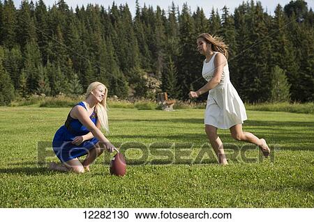 Stock Image - Two girlfriends playing football in a park in their dresses; Edmonton, Alberta, Canada. Fotosearch