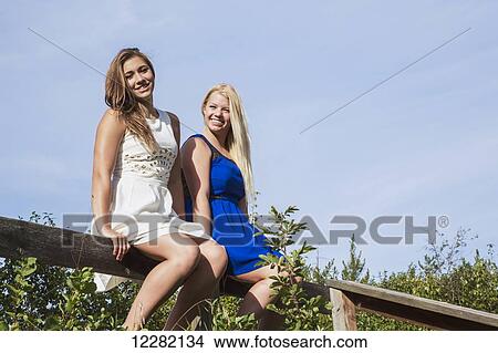 Picture - Two girlfriends sitting on a fence in their dresses in a park; Edmonton, Alberta, Canada. Fotosearch