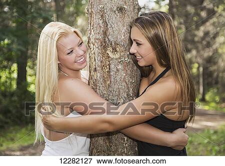 Stock Photo - Two girlfriends smiling at each other while hugging a tree together in a park; Edmonton, Alberta, Canada. Fotosearch