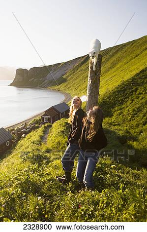Stock Image - Two Girls Watching Their White Kitten Atop A Piling As They Enjoy The Sunset From Their Family's Fish Camp; False Pass, Alaska, United States Of America. Fotosearch