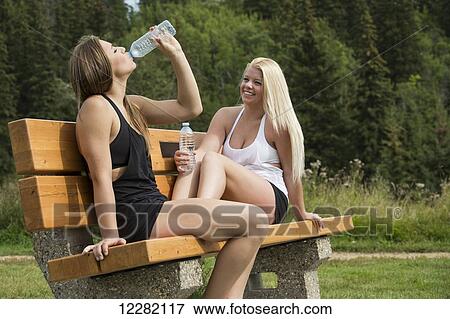 Stock Photo - Two teenage girlfriends sitting on a park bench and drinking bottled water while they rest; Edmonton, Alberta, Canada. Fotosearch