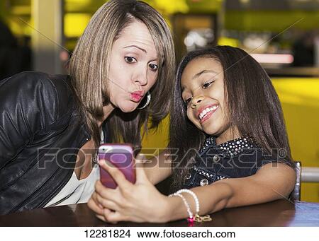 Young mother and her daughter in a cafe looking at a smart phone and taking a selfie while taking a break from shopping; St. Albert, Alberta, Canada View Large Photo Image Picture - Young mother and her daughter in a cafe looking at a smart phone and taking a selfie while taking a break from shopping; St. Albert, Alberta, Canada. Fotosearch