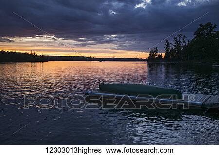 Stock Photograph - A canoe sits on a wooden dock in a tranquil lake at sunset; Ontario, Canada. Fotosearch