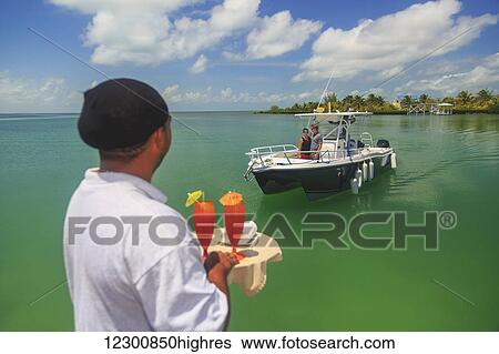 Stock Photograph - A couple in a boat approaches a dock where a server is waiting with drinks, Saint Georges Caye Resort; Belize City, Belize. Fotosearch