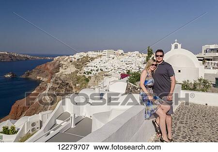 Stock Photo - A couple posing together on a white wall on a greek island with the town in the background; Santorini, Greece. Fotosearch