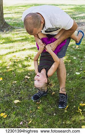 Stock Photograph - A father hanging his daughter upside down and playing around in a park during a family outing; Edmonton, Alberta, Canada. Fotosearch
