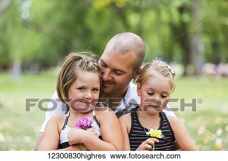 Stock Photograph - A father holding his two daughters in a park; Edmonton, Alberta, Canada. Fotosearch