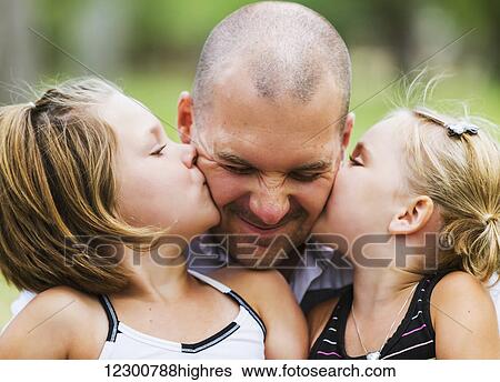 Stock Photograph - A father holding his two daughters who are kissing him in a park; Edmonton, Alberta, Canada. Fotosearch