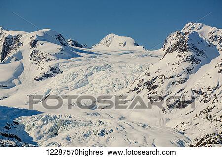 Stock Photograph - A glacier hangs in a valley below jagged snow covered peaks in Kings Bay, Prince William Sound; Whittier, Alaska, United States of America. Fotosearch