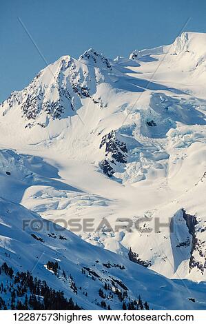 Stock Photograph - A glacier hangs in a valley below jagged snow covered peaks in Kings Bay, Prince William Sound; Whittier, Alaska, United States of America. Fotosearch