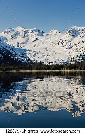Stock Photograph - A glacier hangs in a valley below jagged snow covered peaks in Kings Bay, Prince William Sound; Whittier, Alaska, United States of America. Fotosearch