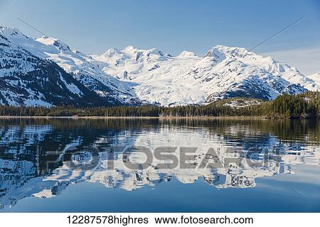 Stock Photograph - A glacier hangs in a valley below jagged snow covered peaks in Kings Bay, Prince William Sound; Whittier, Alaska, United States of America. Fotosearch