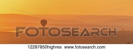 A hot air balloon flies over the African savannah in the orange light before sunrise, with a trail of dust below made by a truck on the ground; Narok, Kenya View Large Photo Image Stock Photograph - A hot air balloon flies over the African savannah in the orange light before sunrise, with a trail of dust below made by a truck on the ground; Narok, Kenya. Fotosearch