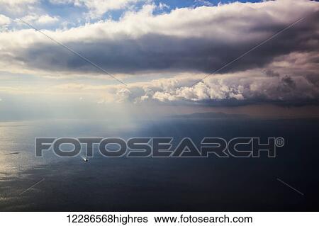 Stock Photograph - A lone boat on the Aegean sea under a cloudy sky and distant view of the coastline; Greece. Fotosearch