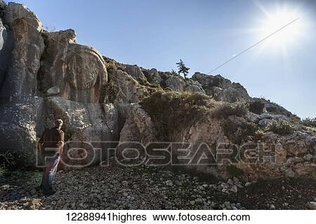 Stock Photograph - A man stands looking at a carving in a rugged rock cliff; Antioch, Turkey. Fotosearch