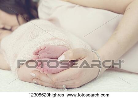 Stock Photograph - A mother laying on a bed with her hands holding a newborn baby's feet; Toronto, Ontario, Canada. Fotosearch