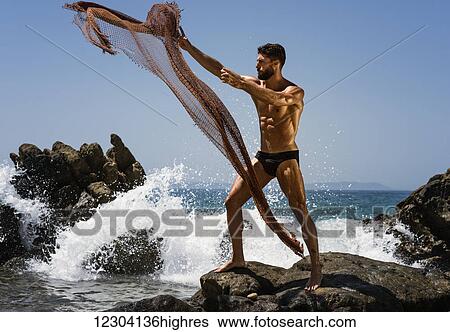 Stock Photograph - A muscular man in a bathing suit throwing a net into the water while standing on a rock at the coast; Tarifa, Cadiz, Andalusia, Spain. Fotosearch