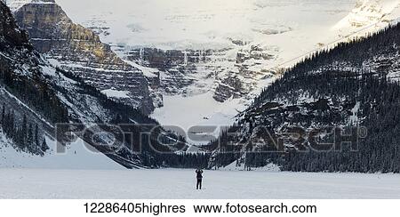 Stock Photograph - A person standing on frozen Lake Louise taking a picture of the snow covered mountains; Lake Louise, Alberta, Canada. Fotosearch