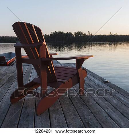 Stock Image - A red adirondack chair sitting on a wooden dock on a lake at sunset; Lake of the Woods, Ontario, Canada. Fotosearch