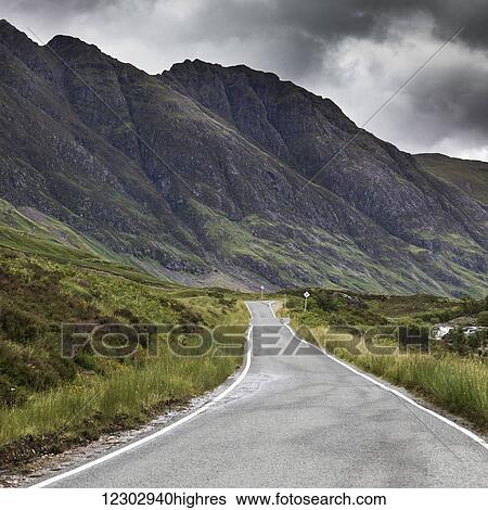 Stock Photograph - A road beside rugged mountains under a stormy sky; Scotland. Fotosearch