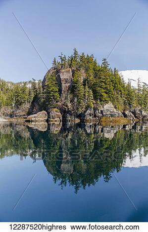 Stock Photograph - A tall outcropping of rocks and evergreen trees reflects on the waters of Prince William Sound; Whittier, Alaska, United States of America. Fotosearch