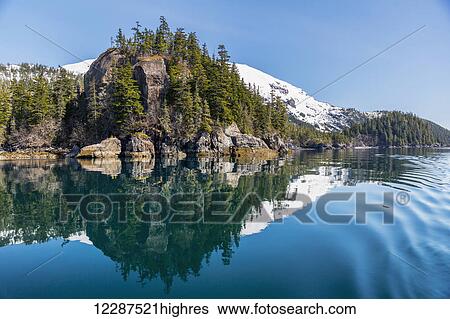 Stock Photograph - A tall outcropping of rocks and evergreen trees reflects on the waters of Prince William Sound; Whittier, Alaska, United States of America. Fotosearch