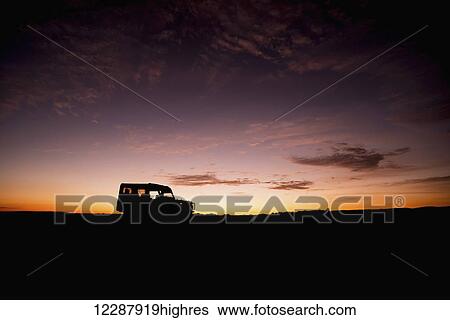 Stock Photograph - A truck parked on the savannah with it's headlights on is shown in silhouette on the horizon at sunrise, blue and orange sky with a few thin clouds in the background; Narok, Kenya. Fotosearch