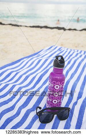 Stock Photograph - A water bottle and sunglasses sit on a beach towel on the sand with view of swimmers in the ocean; Oranjestad, Aruba. Fotosearch