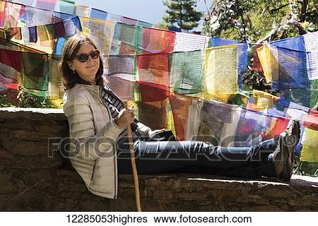 Stock Photograph - A woman rests on a wall beside colourful prayer flags; Paro, Bhutan. Fotosearch