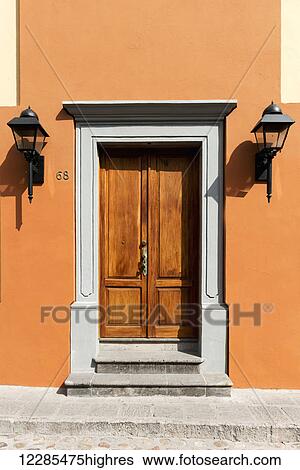 Stock Photograph - A wooden door with white frame and orange walls and two lights on either side; San Miguel de Allende, Guanajuato, Mexico. Fotosearch