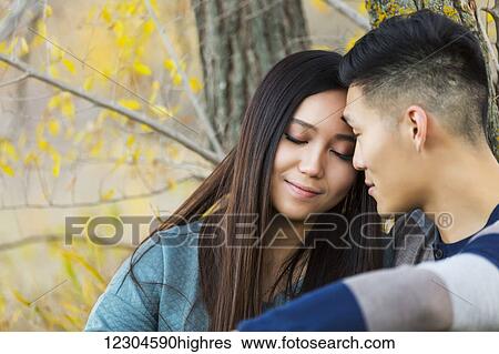 Stock Photograph - A young Asian couple enjoying a romantic time together and sitting down under a tree in a park in autumn; Edmonton, Alberta, Canada. Fotosearch