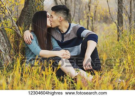 A young Asian couple enjoying a romantic time together outdoors in a park in autumn and kissing each other while sitting under a tree; Edmonton, Alberta, Canada View Large Photo Image Stock Photograph - A young Asian couple enjoying a romantic time together outdoors in a park in autumn and kissing each other while sitting under a tree; Edmonton, Alberta, Canada. Fotosearch