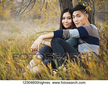 Stock Photograph - A young Asian couple enjoying a romantic time together outdoors in a park in autumn sitting under a tree and posing for the camera; Edmonton, Alberta, Canada. Fotosearch