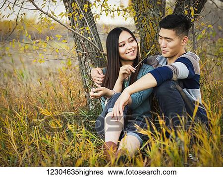 Stock Photograph - A young Asian couple enjoying a romantic time together and sitting under a tree in a park in autumn and she is tickling his nose with a blade of grass; Edmonton, Alberta, Canada. Fotosearch