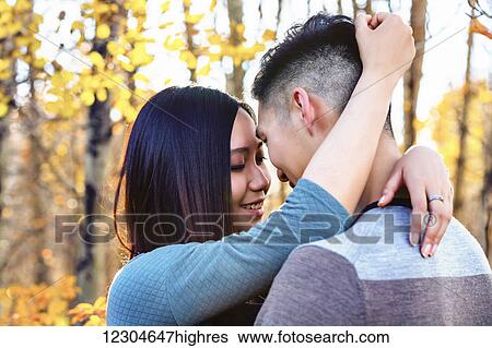 Stock Photograph - A young Asian couple enjoying a romantic time together outdoors in a park in autumn and embracing each other in the warmth of the sunlight during the early evening; Edmonton, Alberta, Canada. Fotosearch