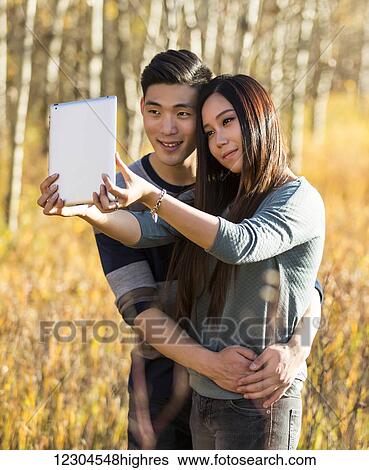 A young Asian couple enjoying quality time together outdoors in a park in autumn and taking self-portraits using a tablet in the warmth of the sunlight during the early evening; Edmonton, Alberta, Canada View Large Photo Image Stock Photograph - A young Asian couple enjoying quality time together outdoors in a park in autumn and taking self-portraits using a tablet in the warmth of the sunlight during the early evening; Edmonton, Alberta, Canada. Fotosearch