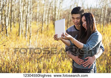 Stock Photograph - A young Asian couple enjoying quality time together outdoors in a park in autumn and taking self-portraits using a tablet in the warmth of the sunlight during the early evening; Edmonton, Alberta, Canada. Fotosearch