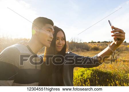 Stock Photograph - A young Asian couple enjoying quality time together outdoors in a park in autumn and taking self-portraits using a smart phone in the late afternoon sunshine; Edmonton, Alberta, Canada. Fotosearch