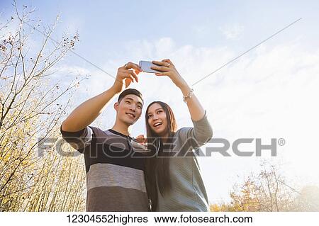 Stock Photograph - A young Asian couple enjoying quality time together outdoors in a park in autumn and taking self-portraits using a smart phone in the late afternoon sunshine; Edmonton, Alberta, Canada. Fotosearch
