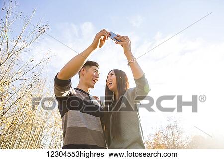 A young Asian couple enjoying quality time together outdoors in a park in autumn and taking self-portraits using a smart phone in the late afternoon sunshine; Edmonton, Alberta, Canada View Large Photo Image Stock Photograph - A young Asian couple enjoying quality time together outdoors in a park in autumn and taking self-portraits using a smart phone in the late afternoon sunshine; Edmonton, Alberta, Canada. Fotosearch