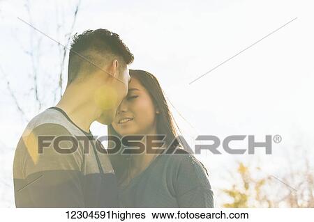 Stock Photograph - A young Asian couple enjoying quality time together outdoors in a park in autumn and touching their foreheads together affectionately in the warmth of the sunlight during the early evening; Edmonton, Alberta, Canada. Fotosearch