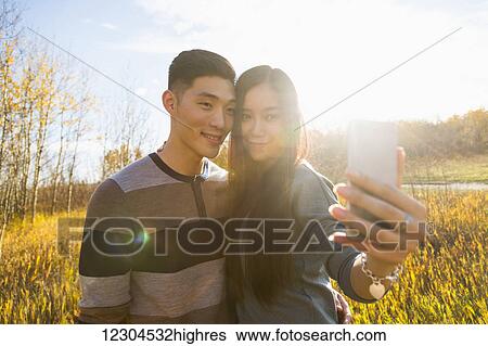 Stock Photograph - A young Asian couple enjoying time together in a park in autumn and taking self-portraits using a smart phone in the late afternoon sunshine; Edmonton, Alberta, Canada. Fotosearch