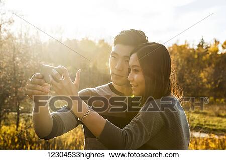 Stock Photograph - A young Asian couple enjoying time together in a park in autumn and taking self-portraits using a smart phone in the late afternoon sunshine; Edmonton, Alberta, Canada. Fotosearch