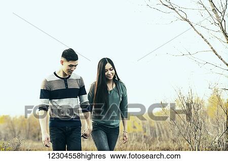Stock Photograph - A young Asian couple holding hands while walking in a park in autumn and and enjoying the warmth of the evening sunshine; Edmonton, Alberta, Canada. Fotosearch