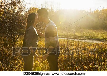 Stock Photograph - A young Asian couple kissing in a park in autumn and making a heart with their hands in the warmth of the setting sun; Edmonton, Alberta, Canada. Fotosearch