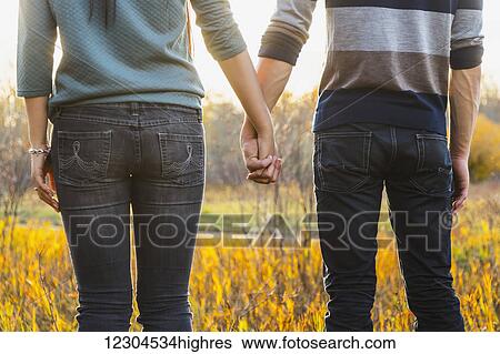 Stock Photograph - A young couple enjoying quality time together outdoors in a park in autumn and holding hands in the warmth of the sunlight during the early evening; Edmonton, Alberta, Canada. Fotosearch