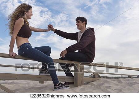 A young man and woman sit talking and holding hands on a wooden fence at the beach; Tarifa, Cadiz, Andalusia, Spain View Large Photo Image Stock Photograph - A young man and woman sit talking and holding hands on a wooden fence at the beach; Tarifa, Cadiz, Andalusia, Spain. Fotosearch