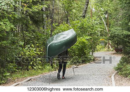 Stock Photograph - A young woman carrying a green canoe on her head down a trail; Ontario, Canada. Fotosearch